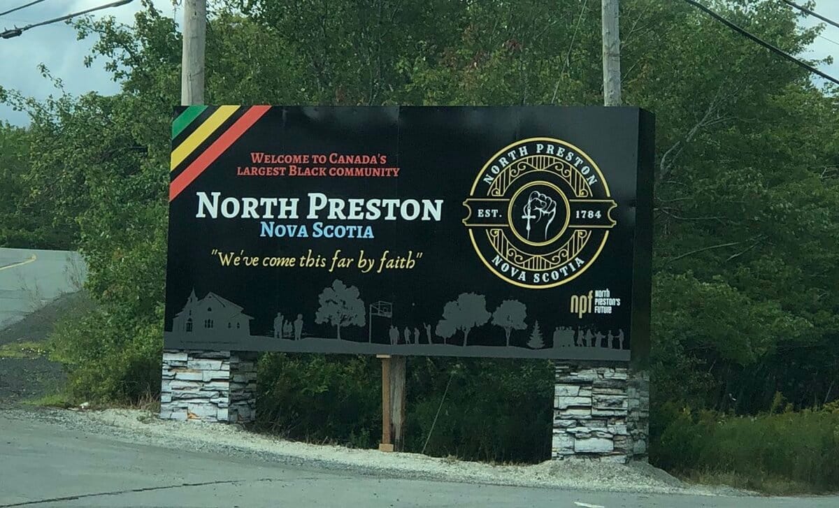 A black road sign with the text "Welcome to Canada's Largest Black Community, North Preston, Nova Scotia" followed by "We've come this far by faith"