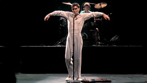 selects from Talking Heads' concert film Stop Making Sense. images of Byrne holding a microphone to camera, raising his arms, dancing in a big suit an with a lamp and full shots of the band.