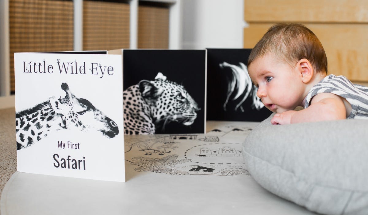 A baby lies on a play mat, focused on a colorful children's book titled "My First Safari" featuring animal illustrations.