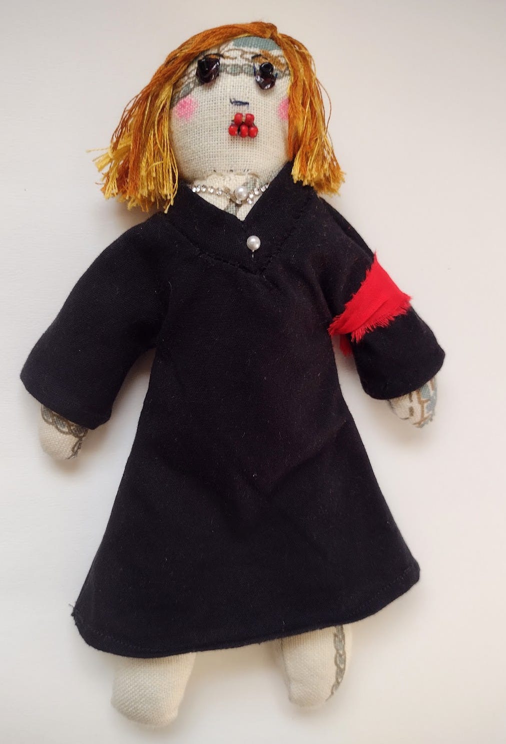 Fabric doll with dirty blonde shoulder length hair, a rhinestone and pearl necklace, black judge's robe, and a red armband