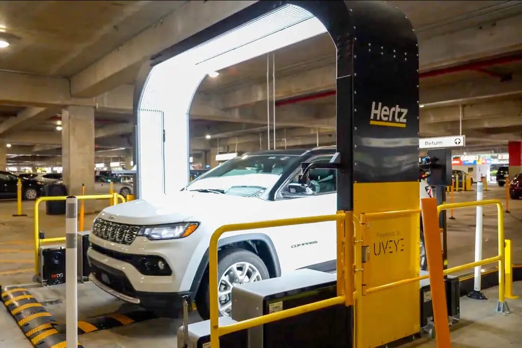 A white Jeep S.U.V. parked in a black and yellow scanner shaped like an upside-down U. The words “Hertz” and “UVeye” are painted on the side of the scanner.