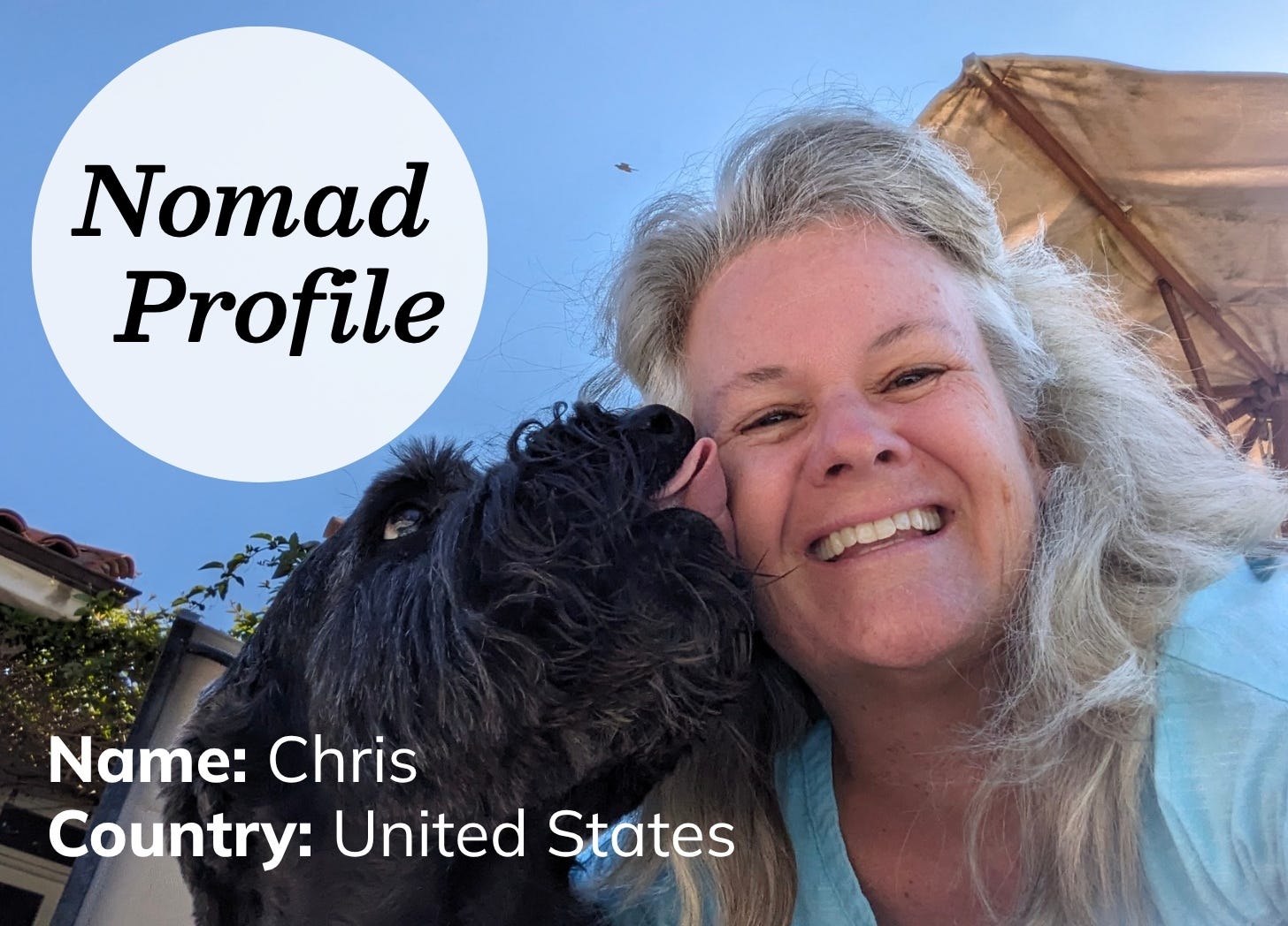 White woman with grey hair is wearing a light blue shirt and smiling. A furry black dog is licking her face. It is a sunny day with blue sky.