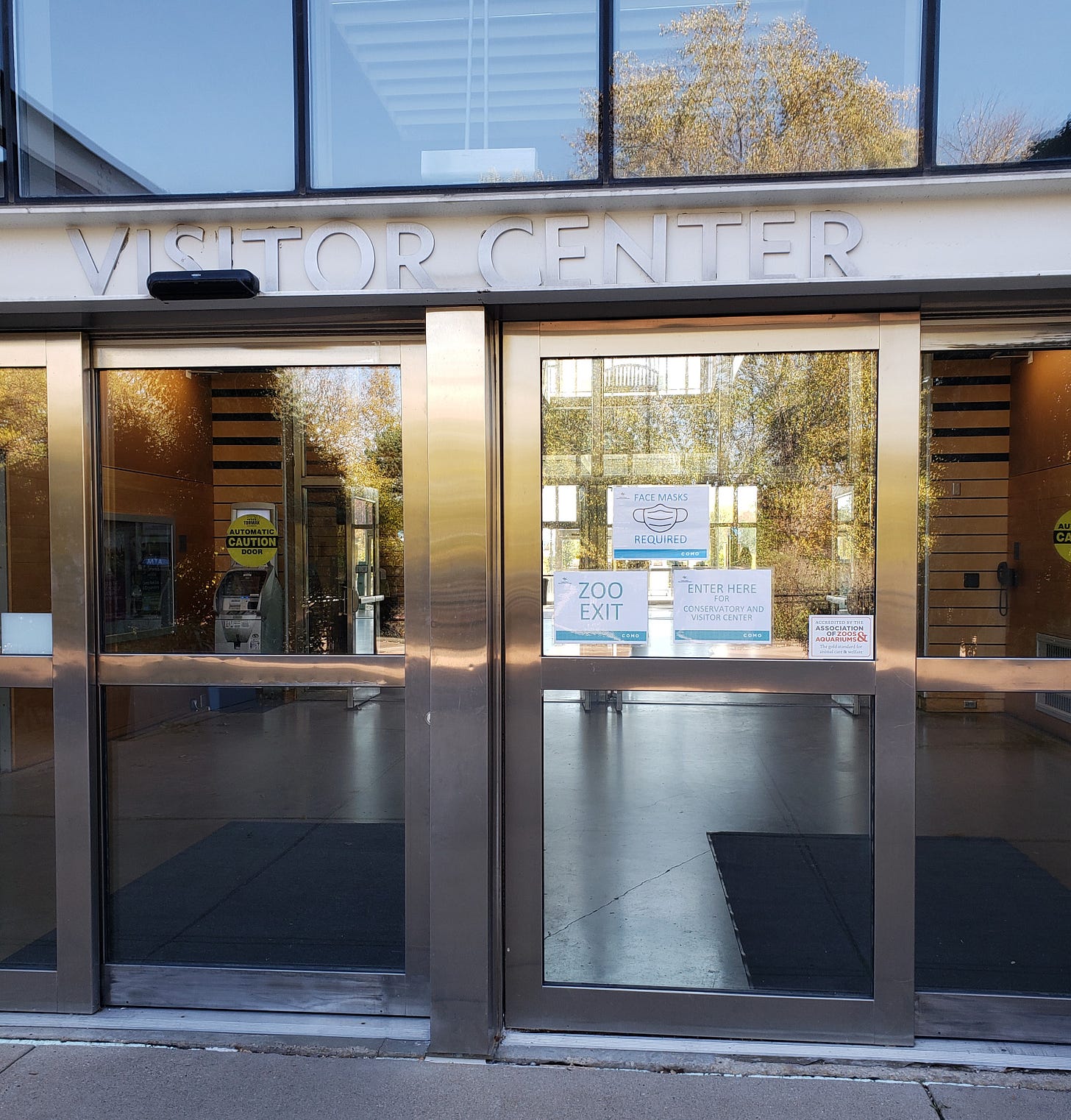 The glass doors of the Visitor Center at Como. There are signs on the door that read Face Masks Required. Zoo Exit. Enter Here for Conservatory and Visitor Center.