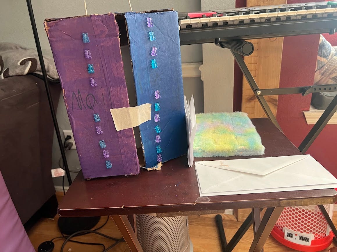 Photo of a cardboard box painted purple and blue with small plastic bears glued in two verticle lines on the front and a piece of tape holding the flaps shut. The box is on a tv tray alongside a fuzzy notebook and a pile of envelopes.
