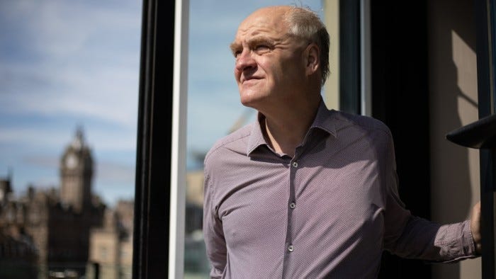 James Anderson stands by an open window, looking out with a thoughtful expression and sunlight on his face.