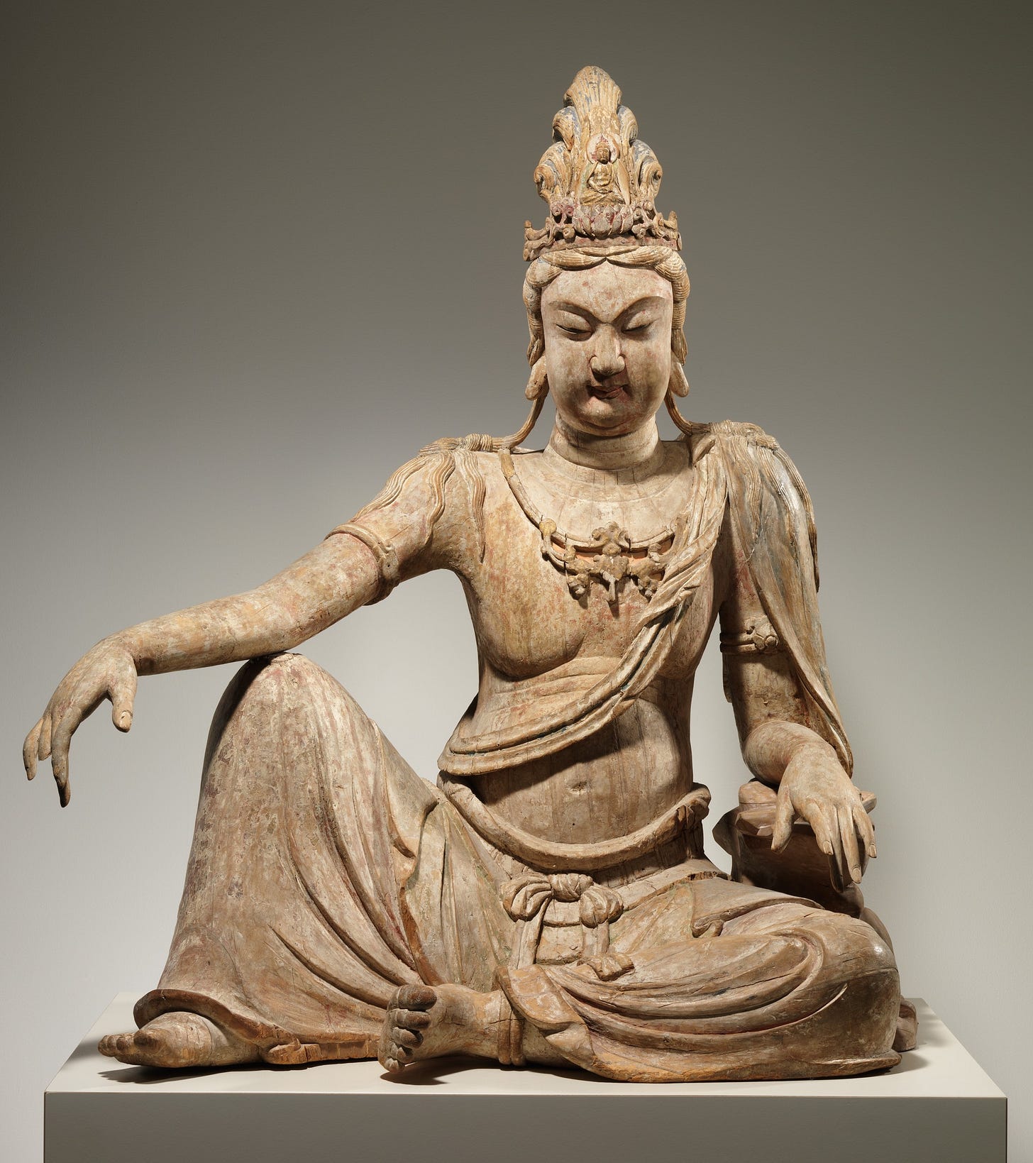 A large wooden sculpture of the bodhisattva Avalokiteshvara or Guanyin in “water moon” pose, with the right knee raised and the left leg crossed before the body. The figure has rounded cheeks, a lowered gaze, and an ornate headdress. The figure appears relaxed and at ease. A large wooden sculpture of the bodhisattva Avalokiteshvara or Guanyin in “water moon” pose, with the right knee raised and the left leg crossed before the body. The figure has rounded cheeks, a lowered gaze, and an ornate headdress. The figure appears relaxed and at ease.