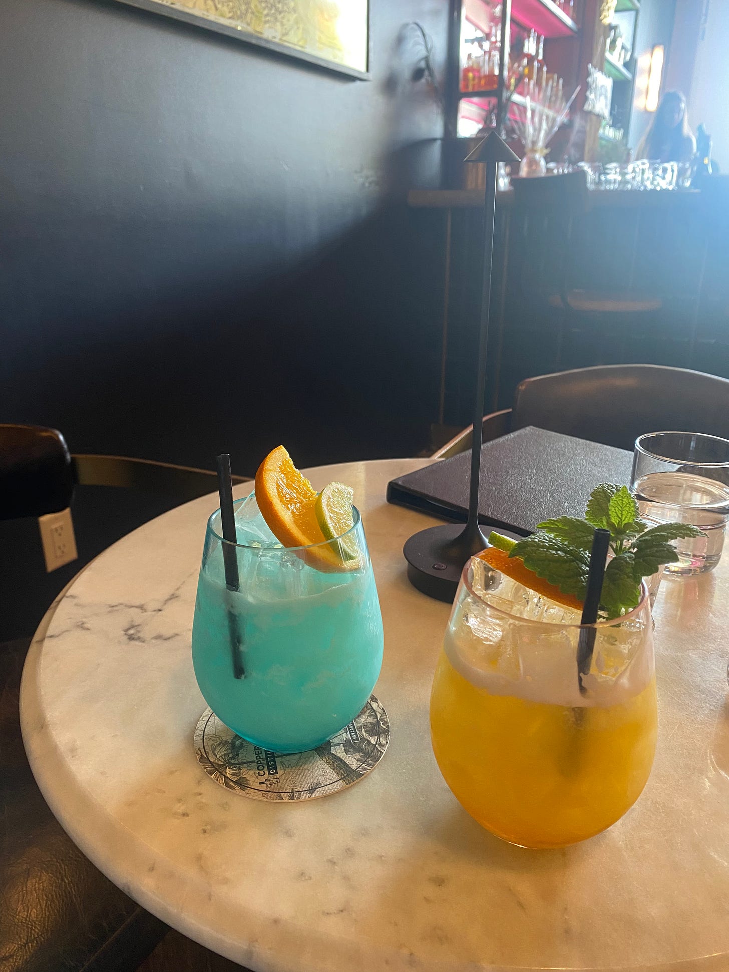 Two drinks in stemless wine-style glasses, one orange and one light blue. Both have ice and fruit garnish. The table is marble and lightly light by the sun filtering in.