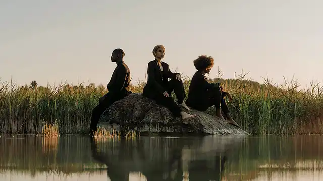 Three people sitting on a rock in tall grass, silhouetted against a pale sky