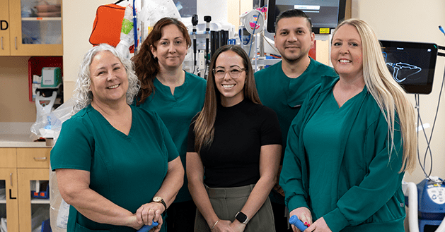 Five Riverside Health Nurses Honored by Virginia Emergency Nurses Association State Council Five smiling healthcare professionals pose in a hospital room, wearing teal scrubs and holding blue medical tools, conveying teamwork.