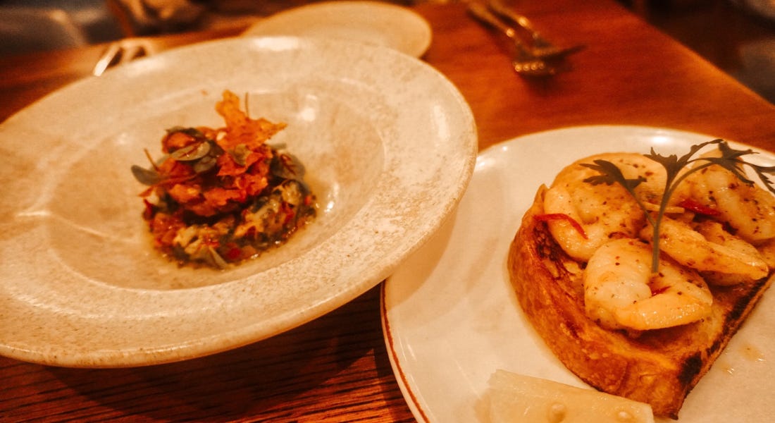Two plates of food - one tartare di tonno and one prawns on toast - on a wooden table Two plates of food - one tartare di tonno and one prawns on toast - on a wooden table