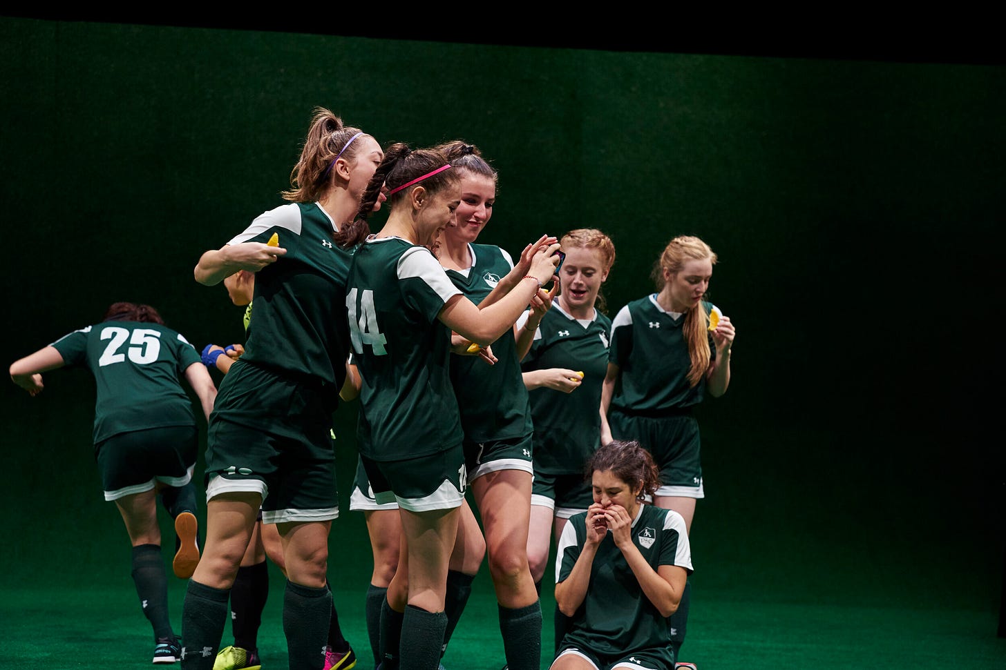 A group of people in green and white soccer uniforms eating orange slices and looking at something together on a cell phone