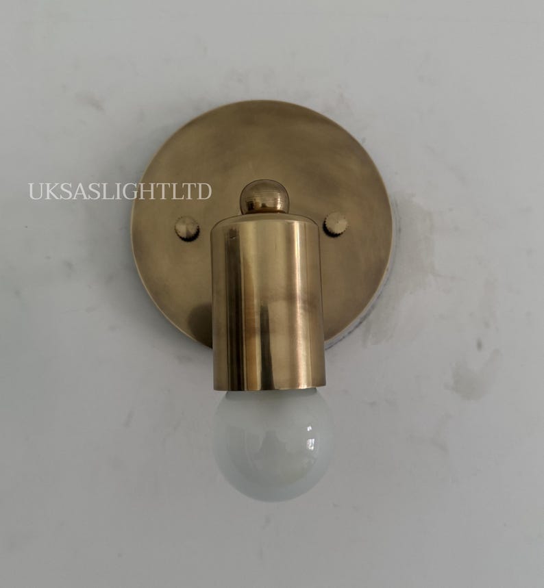May include: A brass-colored wall sconce with a round backplate and a cylindrical light fixture. The sconce has a small, round bulb. The text "UKSASLIGHTLTD" is printed on the backplate. May include: A brass-colored wall sconce with a round backplate and a cylindrical light fixture. The sconce has a small, round bulb. The text "UKSASLIGHTLTD" is printed on the backplate.