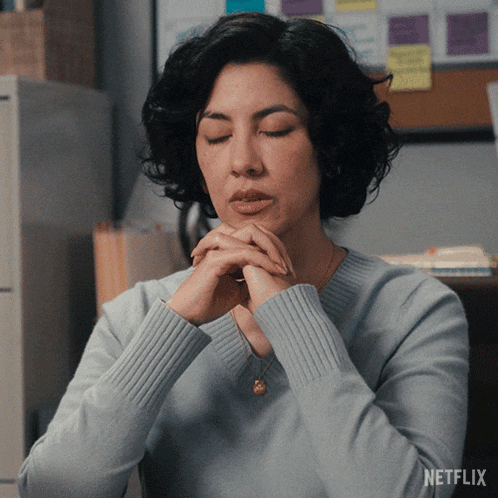 GIF d'une femme aux cheveux bruns bouclés, yeux fermés et mains jointes devant le visage, l'air découragée à son bureau.