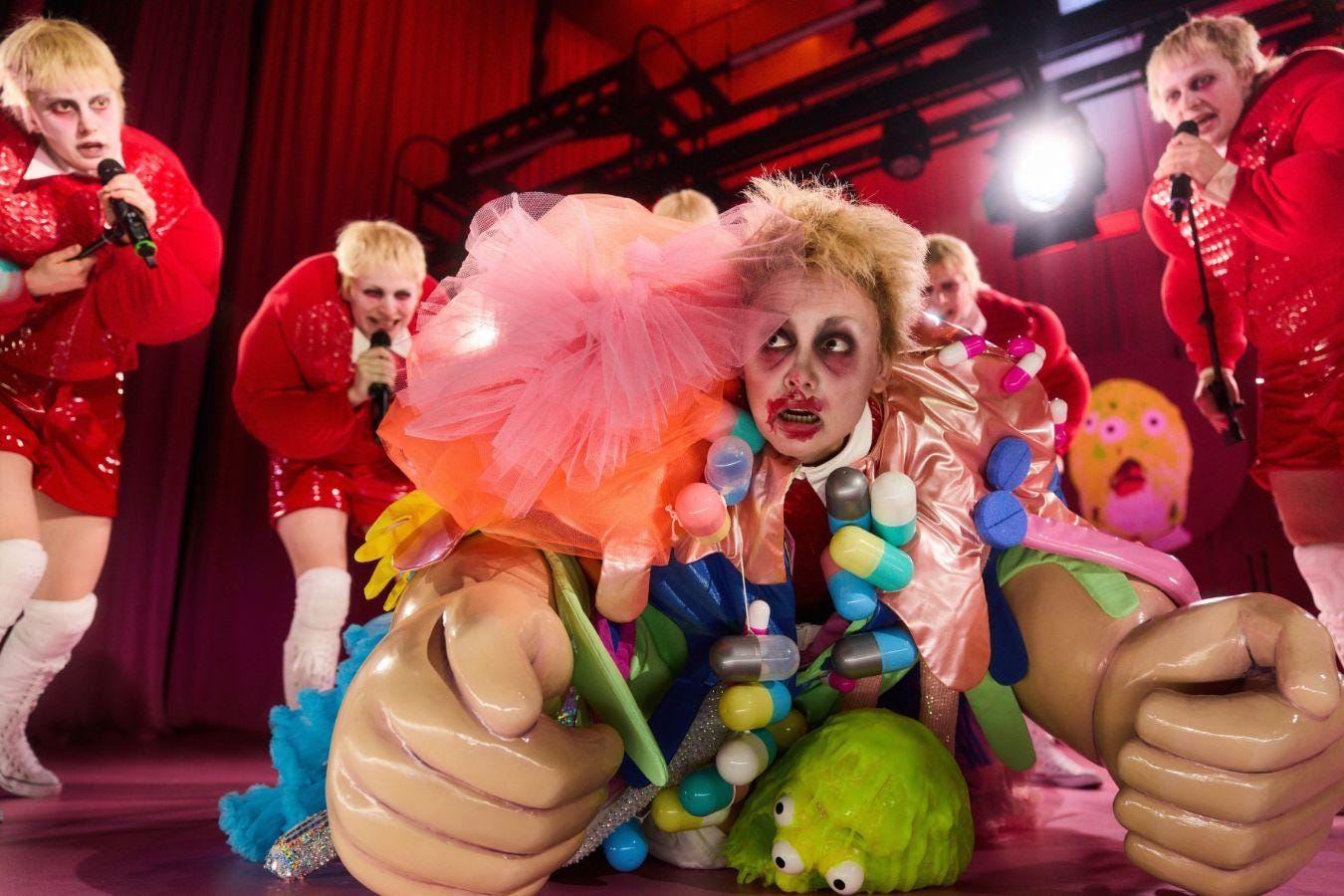 Stage performance with several people in glittering red costumes; in the foreground, a brightly costumed figure with large oversized prop hands and colourful decorative elements, surrounded by bright stage lighting.