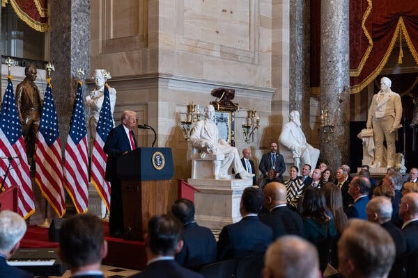 Mr. Trump standing at a lectern and speaking into a microphone in front of an audience. There are statues behind him and to his left.