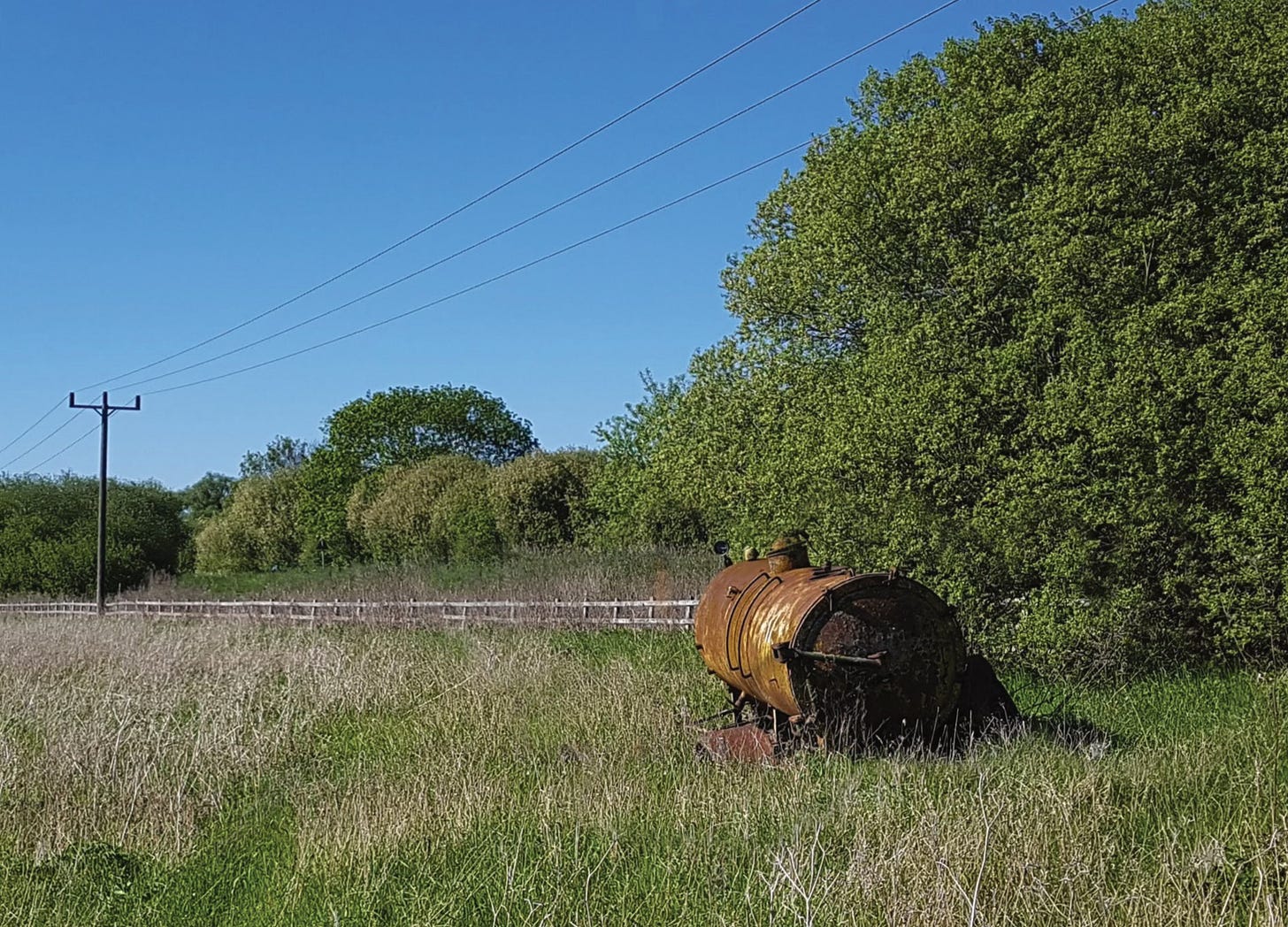 Photograph of a rusty old water bowser in a grassy field on a sunny day with blue sky