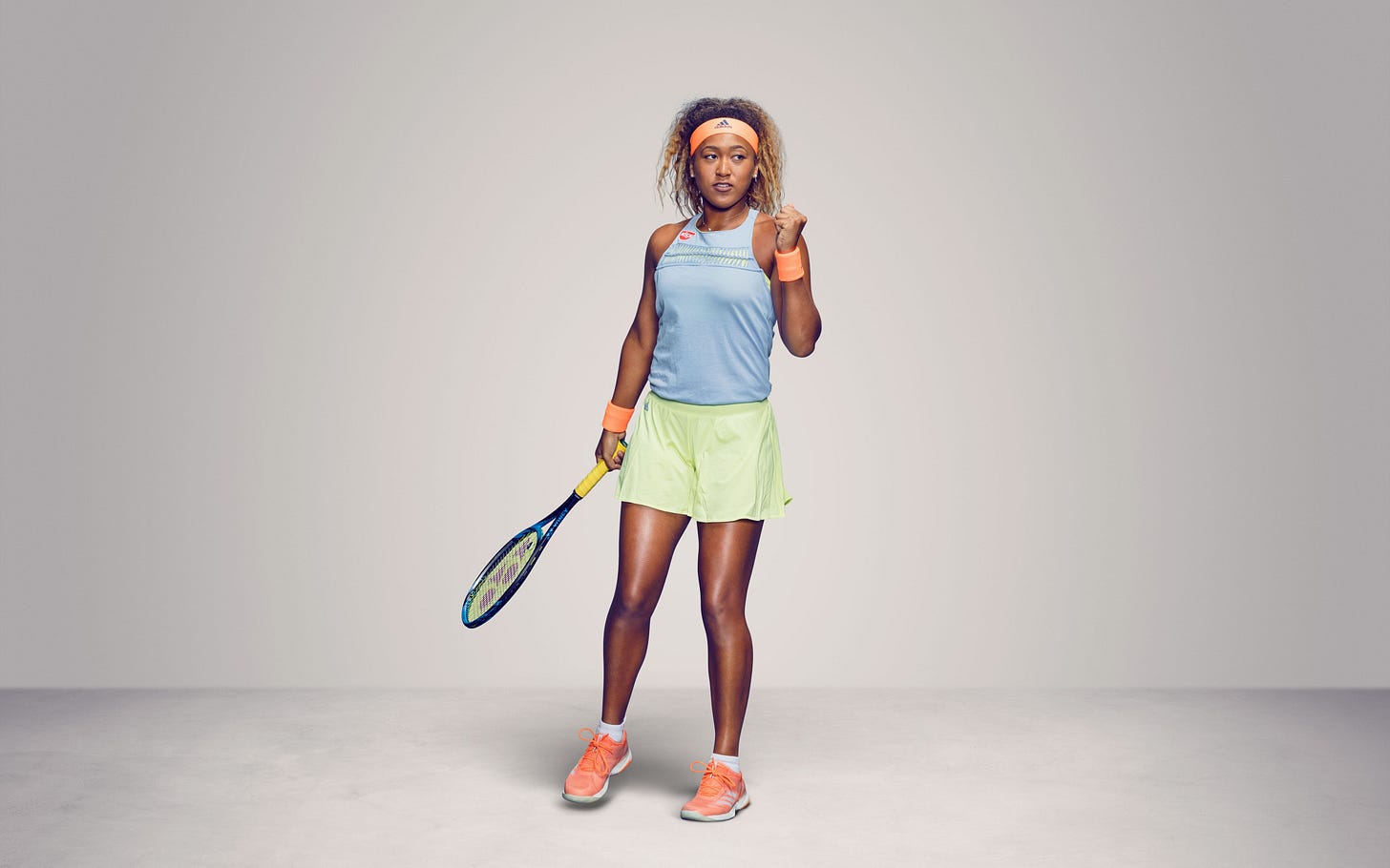 La tennista Naomi Osaka con la racchetta nella mano destra e la mano sinistra alzata a pugno. Indossa una maglietta azzurra e pantaloncini verdi. Ha lo sguardo rivolto fuori campo. Dietro di lei, uno sfondo grigio.