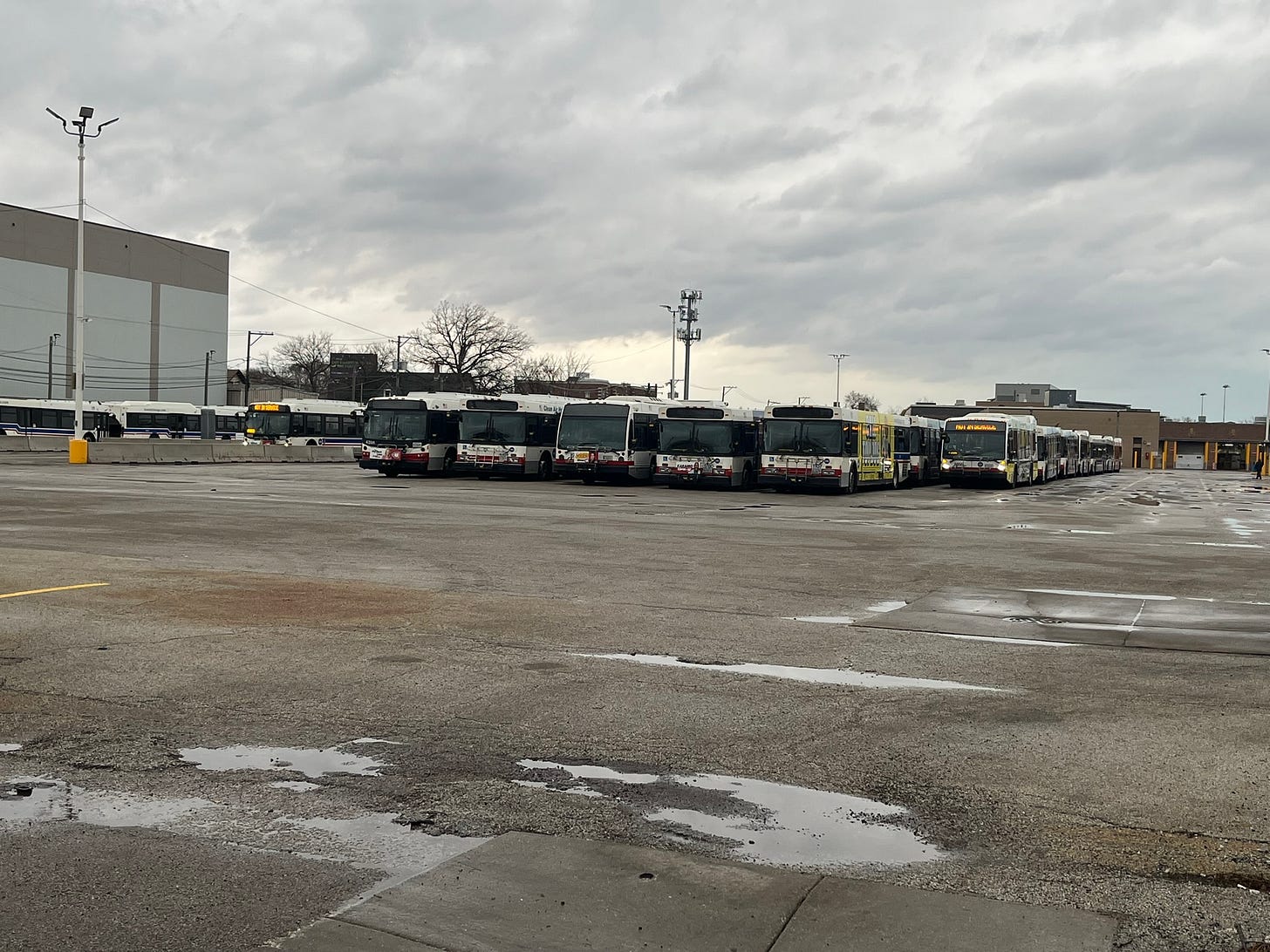 6 rows of buses parked in a large parking lot, with more parked buses to the left, beyond a concrete barrier 6 rows of buses parked in a large parking lot, with more parked buses to the left, beyond a concrete barrier