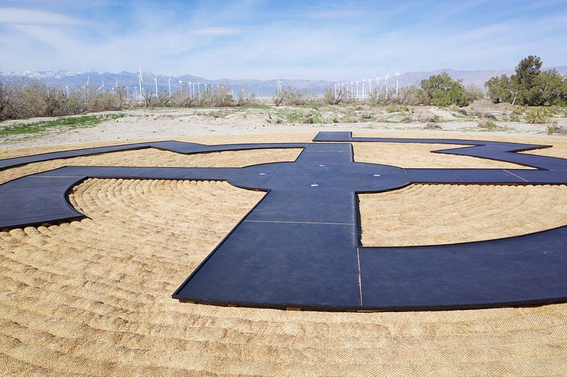 Rows of wind turbines are of in the distance. In the foreground is a large, woven sculpture. Coils of basket-weaving material form a flat disc. On top of this is a black, maze-like pathway. Rows of wind turbines are of in the distance. In the foreground is a large, woven sculpture. Coils of basket-weaving material form a flat disc. On top of this is a black, maze-like pathway.