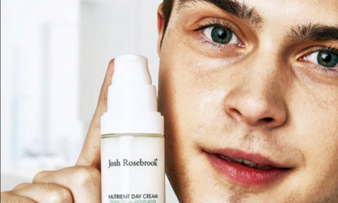 a model with green eyes peers close to the camera and holds a bottle of josh rosebrook nutrient day cream next to their face.