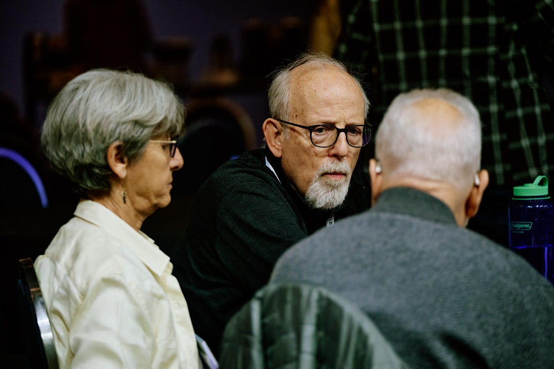 Older man in black glasses leaning in close to say something quietly to the person beside him, woman in cream blouse to his left