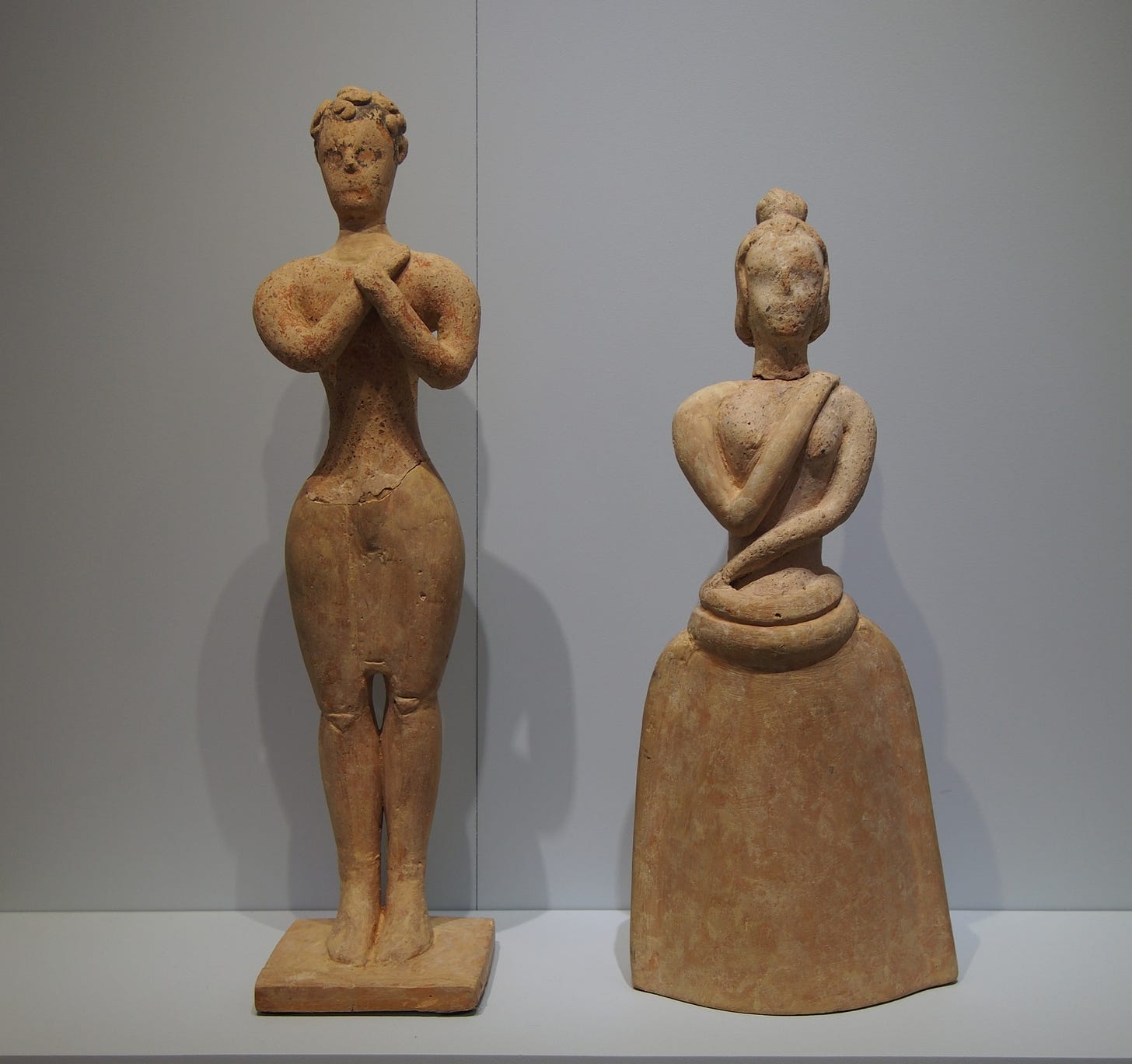 Two tan colored ceramic figurines, one shaped like a man and the other like a woman, stand in a museum exhibit in front of a white wall.