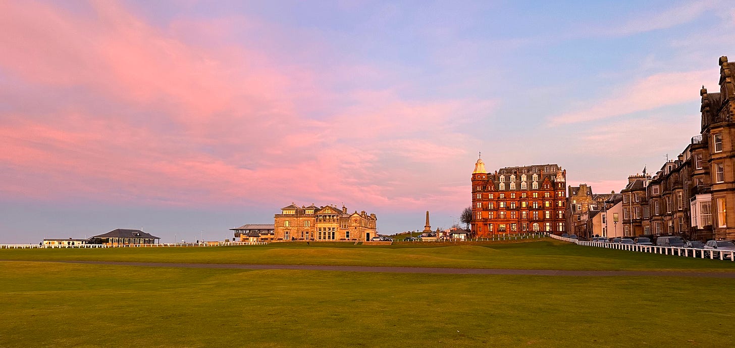 The Old Course at St Andrews beneath a pink evening sky, with the historic clubhouse, hotels and town buildings in the background.