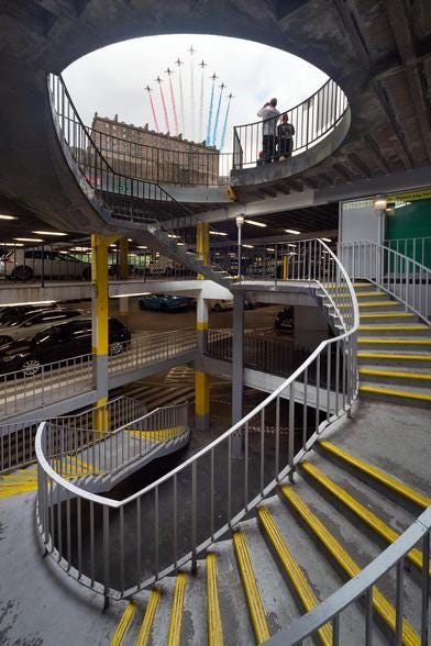 A spiral staircase in a grey, concrete carpark. At the top you can see out to the sky, and in the background is Edinburgh Castle with the Red Arrows flying over. 