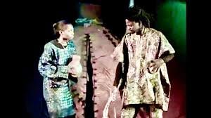 Still from The Palm-Wine Drinkard folk opera. The palm-wine drinkard stands looking at a woman holding a jug of delicious palm-wine.