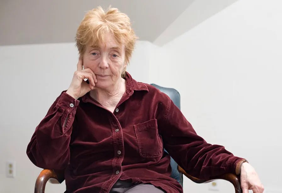 A person with short blonde hair wearing a burgandy shirt. She is sitting in a chair.