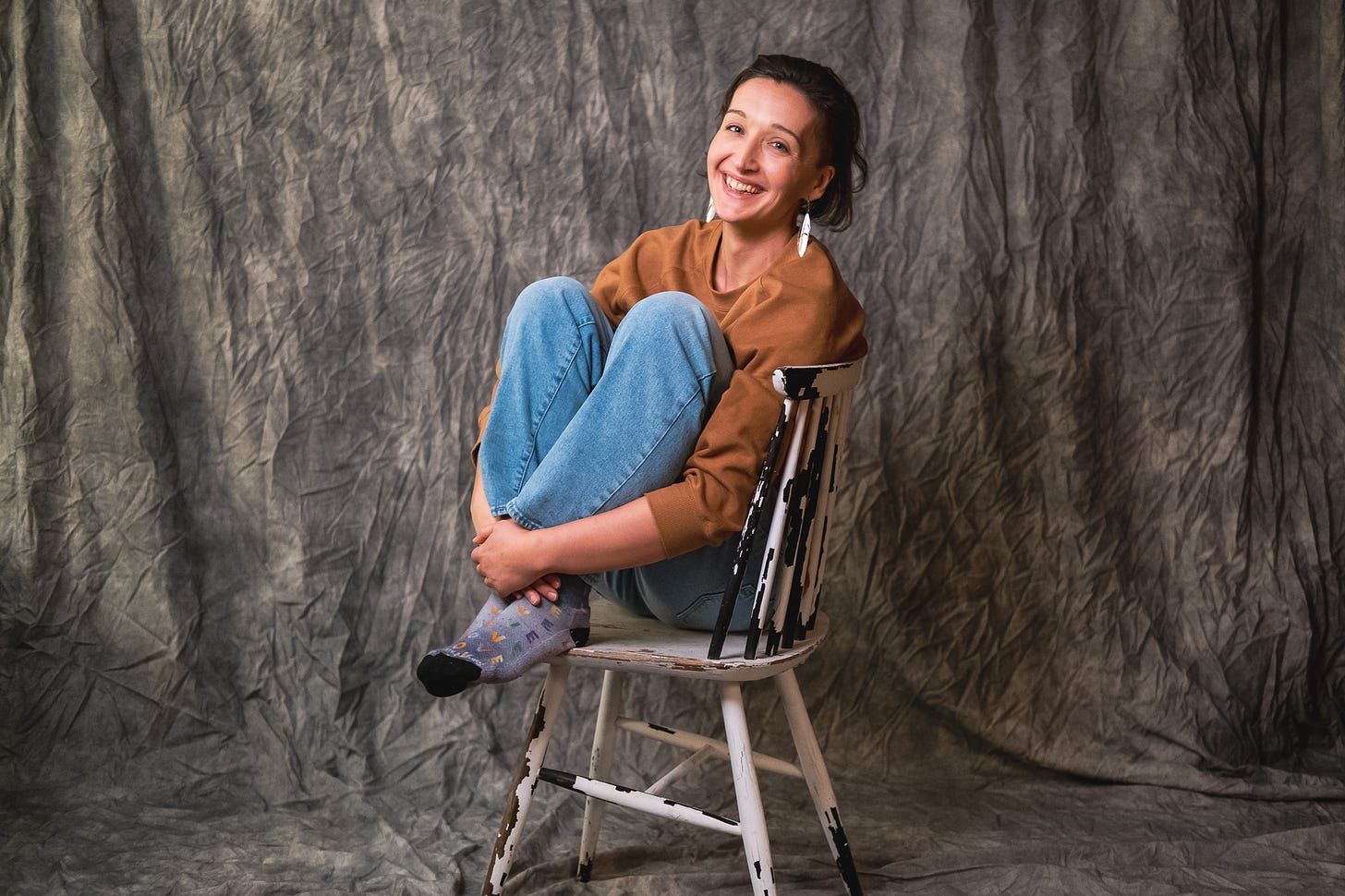 Tarzan sits in a paint-chipped white dining chair with her knees up to her chest, in a comfy mustard sweater and cozy sock, a smile on her face