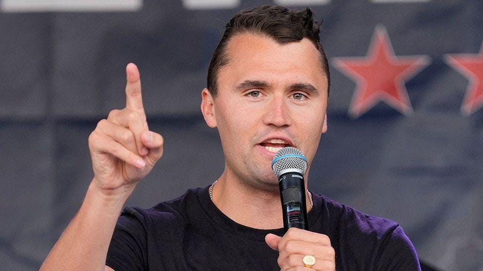 Charlie Kirk speaks into a microphone while holding up his right hand pointing his index finger. He has short dark hair and is wearing a black t-shirt. Charlie Kirk speaks into a microphone while holding up his right hand pointing his index finger. He has short dark hair and is wearing a black t-shirt.