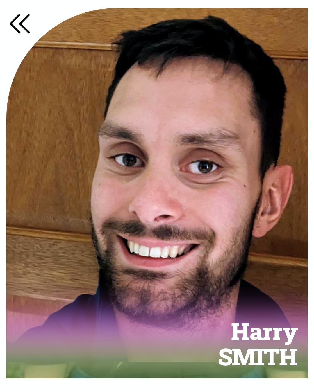 Harry is a young white man with short black hair and a cropped beard. He’s standing against a wooden wall, wearing a navy blue shirt and smiling into the camera. The bottom of the photo bears purple and green streaks, with Harry SMITH written in white.