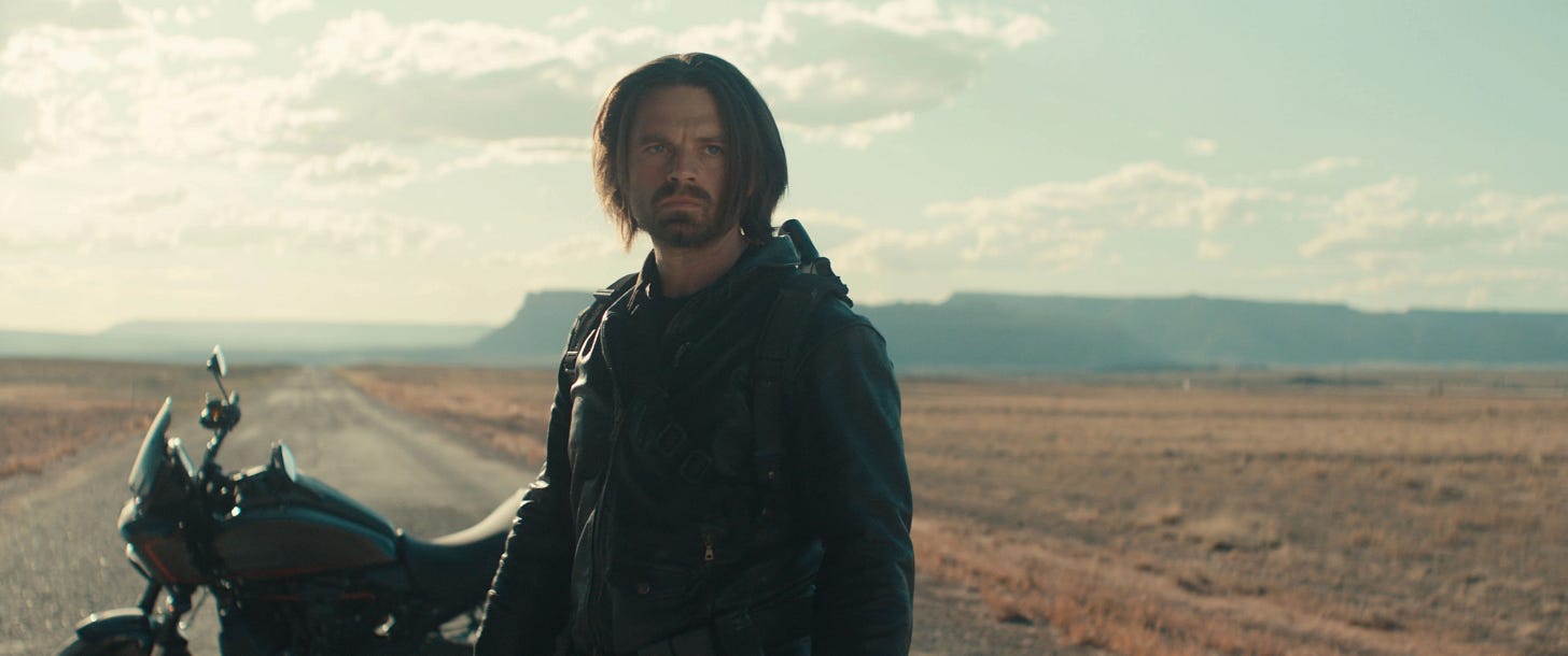 Bucky wearing a leather jacket standing on a desert highway with his motorbike behind him