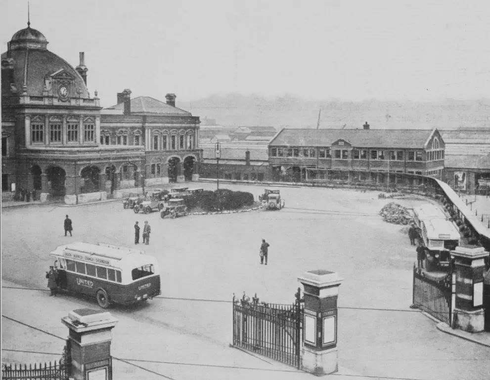 b/w photo of Norwich Station in 1931 – the earliest image I could find. The station is in the background to the left, with a large area in front, enclosed by a low wall with gateposts in the foreground. In this forecourt stand some cars of the period and a bus branded ‘United’