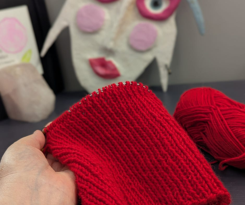 Close-up of a bright red knitted hat in progress, held in hand, showing ribbed stitches and crown shaping, with a ball of red yarn in the background.