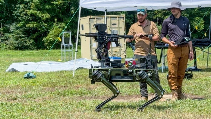 A man videos a robotic four-legged unmanned ground vehicle with a mounted rifle gun standing in the grass with trees in the background.