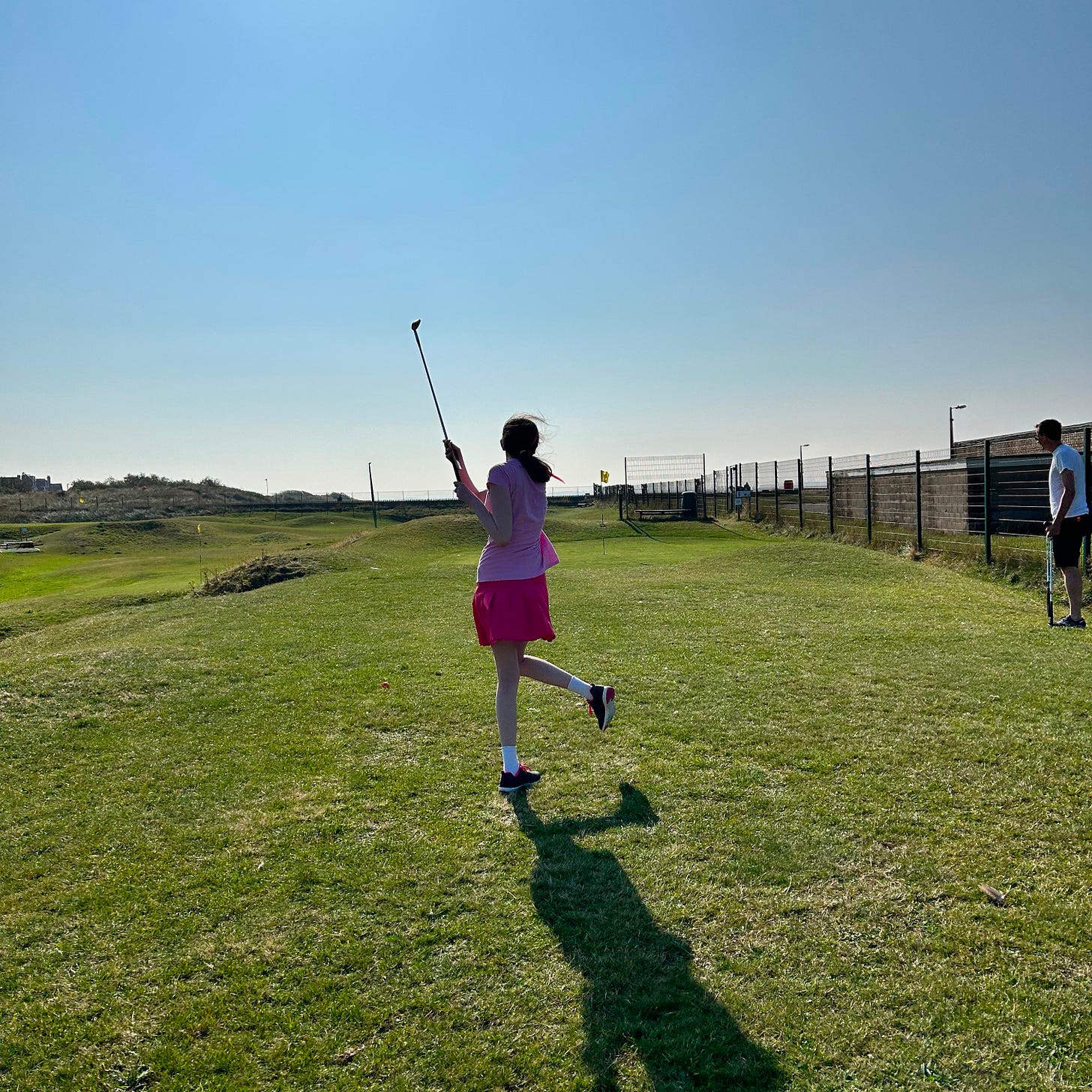 Tall girl in pink shirt and pink skirt playing golf on a course with bright blue skies