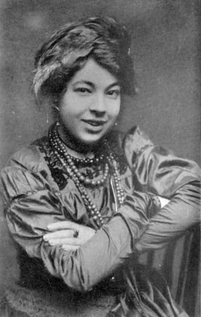 Une jeune femme souriante, l'air un peu mutin, en tenue de la Belle Époque avec force colliers et une jolie coiffe entre la tiare et le turban. 