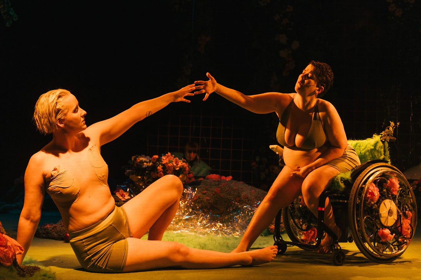 Photo from “Eden.” Embla and Nína, white non-binary actor, on stage reaching towards each other, mimicking the famous painting The Creation of Adam by Michelangelo. Nína is seated on the ground and Embla in her wheelchair. The background shows green grass, pink flowers and off focus is an electronic musician. Photo from “Eden.” Embla and Nína, white non-binary actor, on stage reaching towards each other, mimicking the famous painting The Creation of Adam by Michelangelo. Nína is seated on the ground and Embla in her wheelchair. The background shows green grass, pink flowers and off focus is an electronic musician.