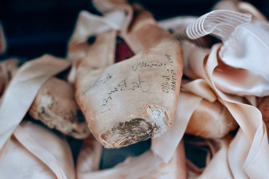 One weathered pink ballet slipper is in focus on a pile of other pink satin pointe shoes. It is signed in black ink by various professional dancers from the Mark Morris Dance Company — including Mark Morris.