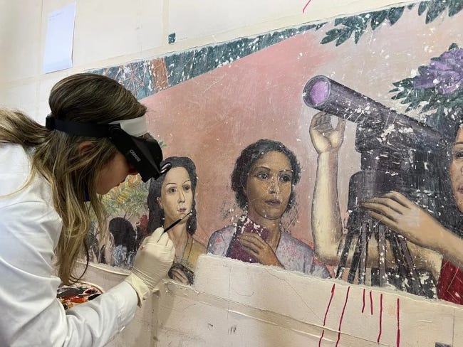 En blouse blanche, lunettes de protection sur les yeux, une femme retouche une partie de la fresque. On voit trois femmes, dont une derrière un téléscope.