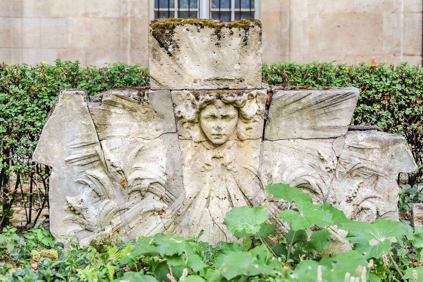 Carved historic stone fragment surrounded by plants in Square Georges Cain, part of the outdoor collection near Musée Carnavalet.