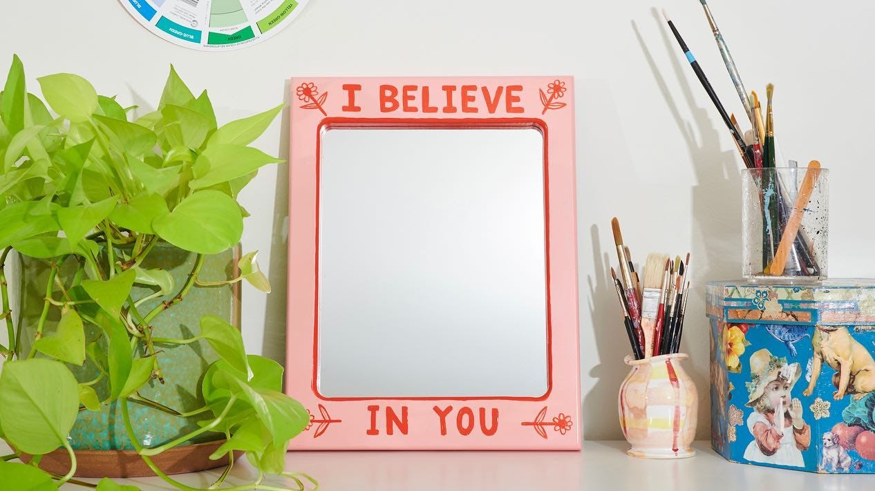 a mirror with a pink frame that contains hand-painted flowers and the words "I believe in you"