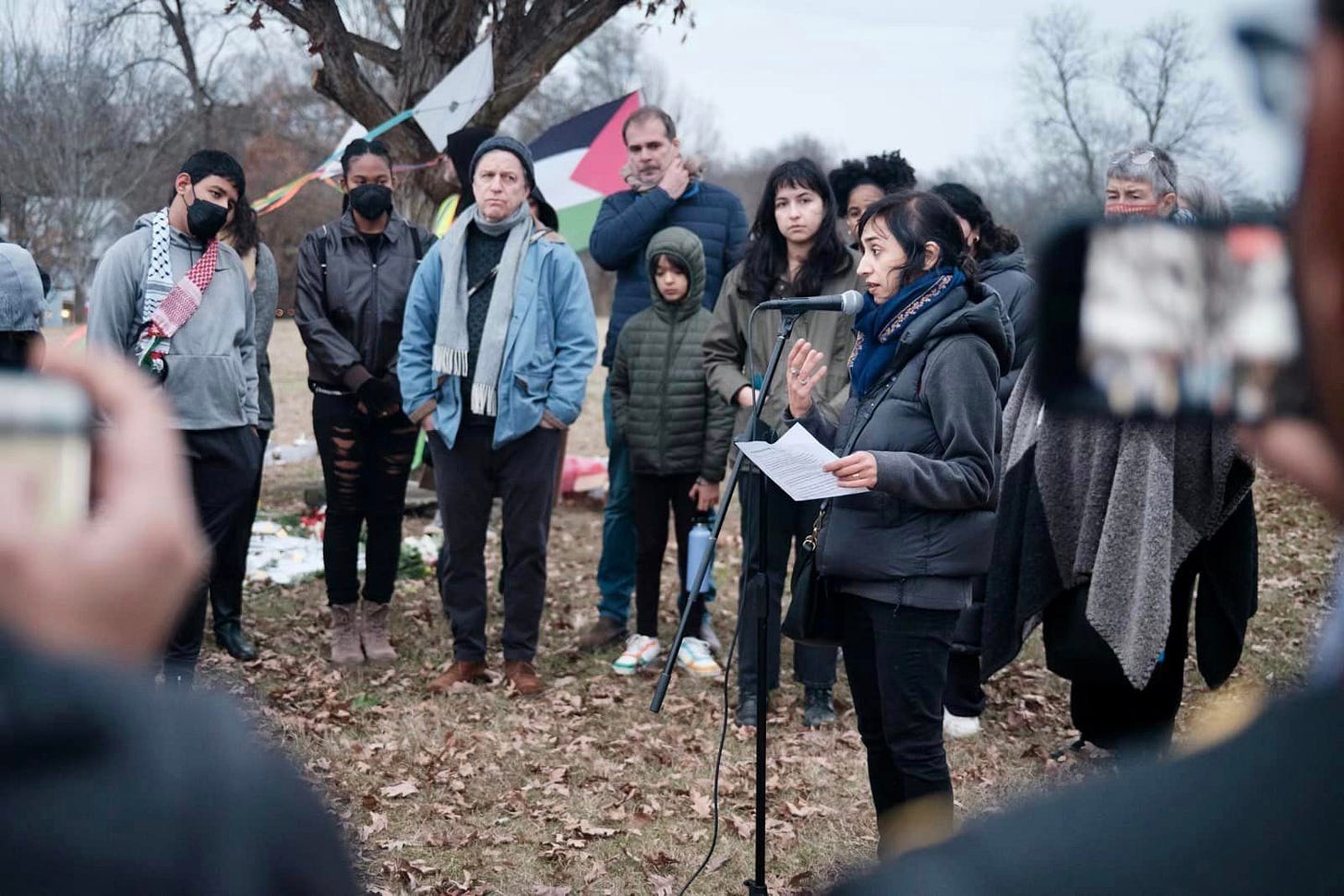 A group of people standing in front of a tree. Sophia Qureshi speaking at a mic. Palestinian flag behind the crowd. 