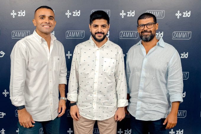 Three men stand side by side in front of a dark blue backdrop branded with Zid and Zammit logos repeated across it. The man on the left wears a white shirt with a subtle pattern, beige pants, and a watch. The middle man is dressed in a white patterned shirt. The right man has on a light blue shirt and beige pants. All appear professional and are smiling at the camera.