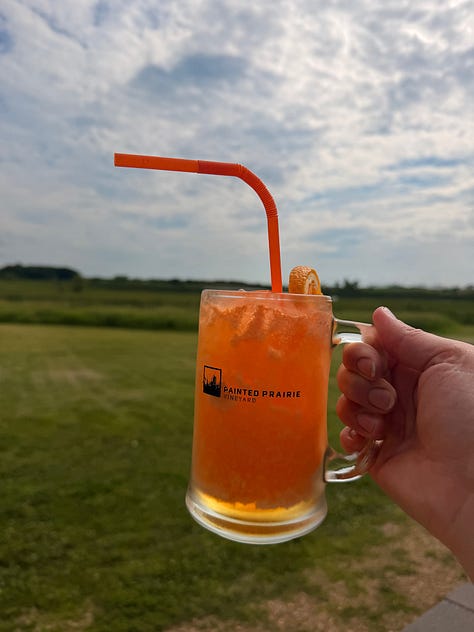 Two very cheesy pizzas on wooden planks and a fizzy drink topped with blackberries in a mug. | A back patio with fire pit, plenty of seating, and a vineyard. | A person holding a bright orange drink in a mug reading “Painted Prairie”
