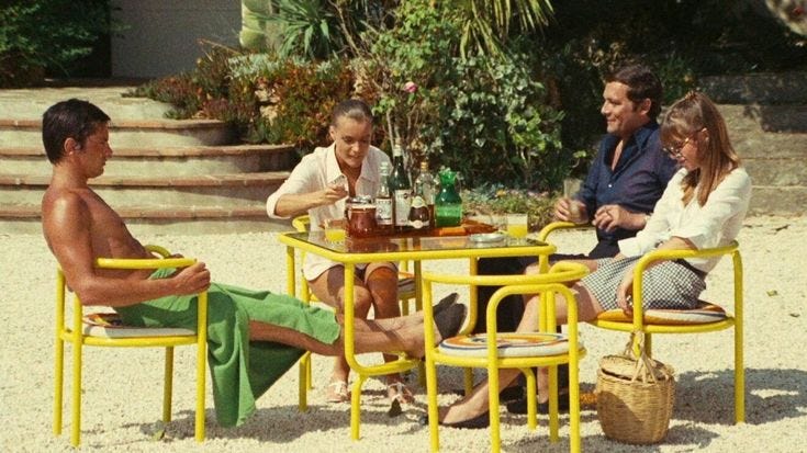 This may contain: four people sitting around a yellow table and chairs in the sand, with one person holding a drink