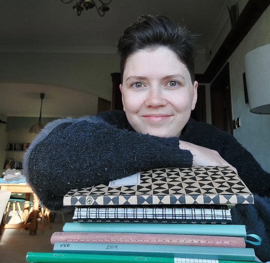 Janelle smiling and looking into the camera. She is a white with very short brown hair in a quiff. She is leaning her arm on a pile of notebooks