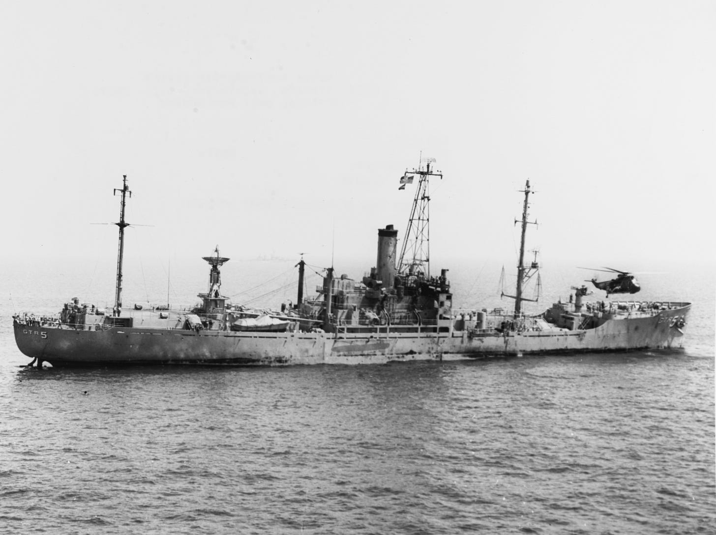A black-and-white photograph of a U.S. Navy ship at sea, labeled “GTR5” on the stern and “AGR5” on the bow, with tall masts and antennas along the deck and a helicopter hovering near the ship’s bow above the water.